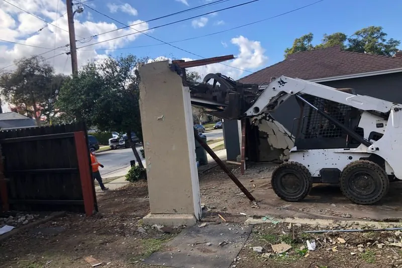 Skid steer loader demolishing a masonry pilaster and perimeter wall at a street frontage